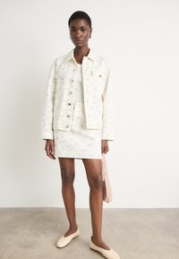 Veste en denim à motif floral et jupe en crème, associées à un haut blanc et des chaussures à enfiler beiges, tenant un sac rose clair.