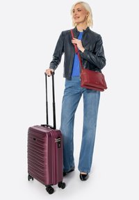 Valigia rigida di colore bordeaux metallico con texture rigata, quattro ruote e manico telescopico, abbinata a una borsa rossa e un abbigliamento casual.