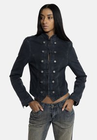 Γυναίκα με μακριά μαύρα μαλλιά που φοράει σκούρο σακάκι με κουμπιά και ξεθωριασμένο γκρι τζιν, όρθια με τα χέρια στις τσέπες, κοιτάζοντας αριστερά.