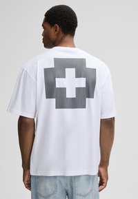 Weißes T-Shirt mit kurzen Ärmeln, das auf der Rückseite eine große schwarze Grafik in Form eines Kreuzes zeigt. Weiches Baumwollmaterial.