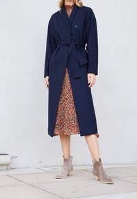Cappotto lungo blu navy con cintura e bottone, abbinato a un vestito floreale e stivaletti taupe alla caviglia. Il cappotto ha un tessuto morbido e testurizzato.