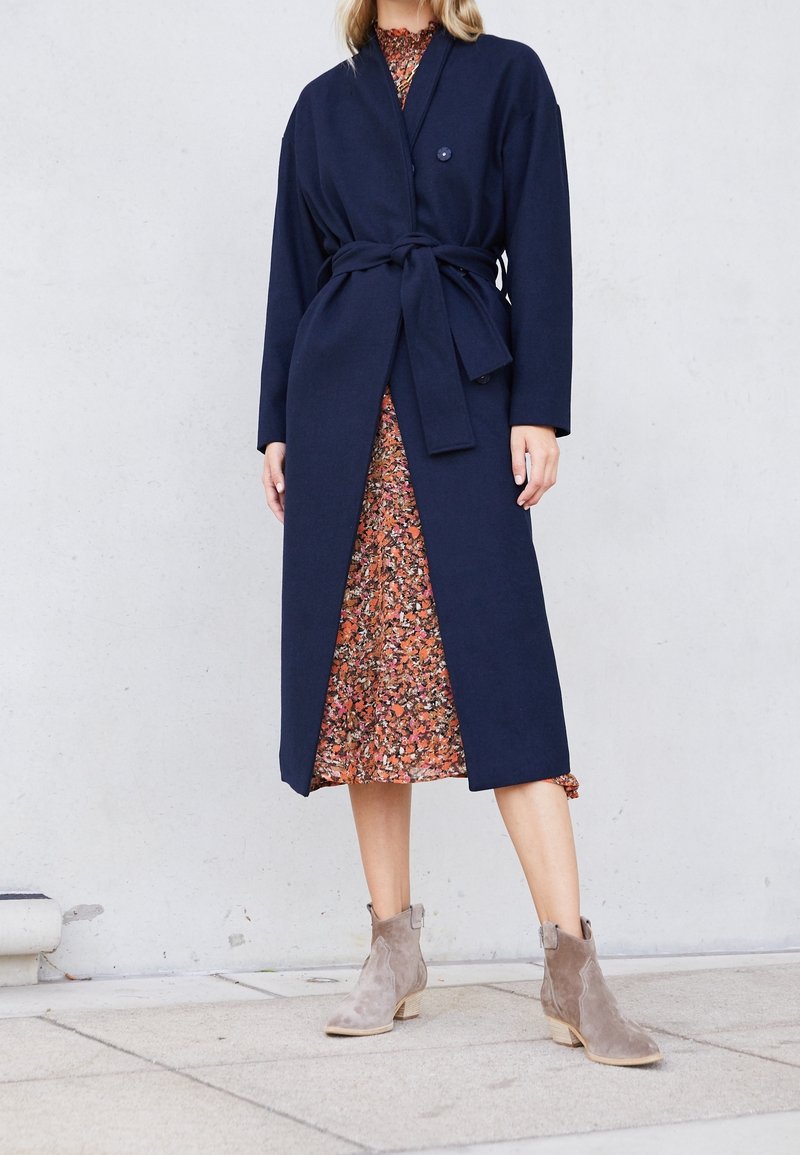 Cappotto lungo blu navy con cintura e bottone, abbinato a un vestito floreale e stivaletti taupe alla caviglia. Il cappotto ha un tessuto morbido e testurizzato.