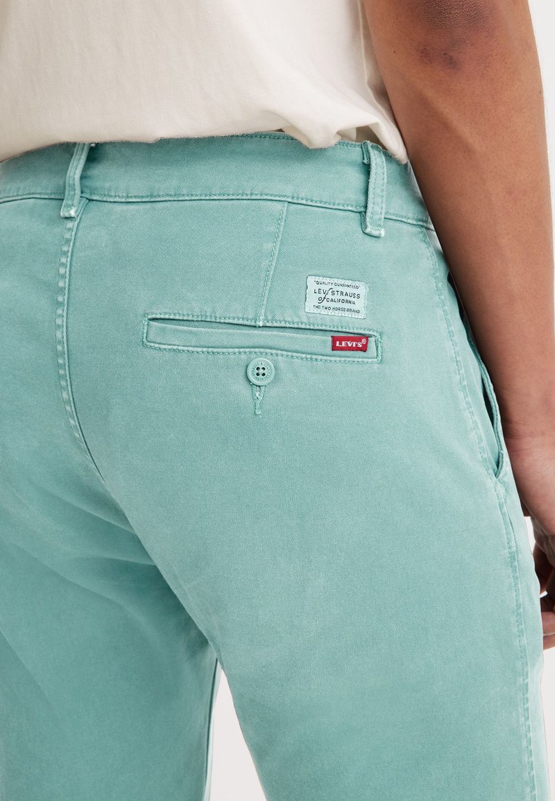 Pantalones de denim verde menta con detalle en el bolsillo trasero, que presentan una etiqueta de Levi's y cierre de botón. Textura suave y diseño ajustado.