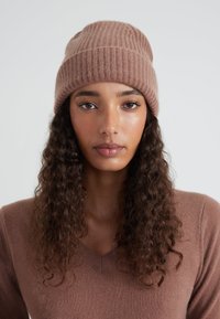 Bruine gebreide muts met een geribbelde textuur en een opgevouwen boord. Het model draagt een bijpassende bruine V-hals trui.