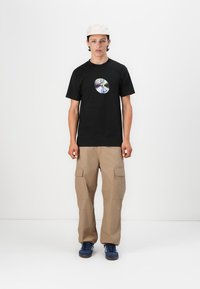 Black t-shirt with a circular graphic on the chest, paired with beige cargo pants. The outfit is completed with blue sneakers and a light cap.