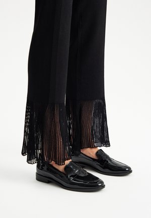 Black ribbed pants with fringed sheer bottoms paired with shiny black loafers feature a decorative bow accent on the front.