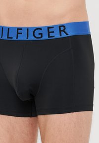 Schwarze Boxer-Shorts mit glatter Textur, ausgestattet mit einem blauen Bund, der mit schwarzen Buchstaben "TOMMY HILFIGER" beschriftet ist.