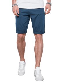 Shorts de algodón azul con puños enrollados, un ajuste relajado y bolsillos laterales, combinados con zapatillas blancas, frente a un fondo liso.