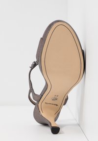 Une sandale grise à talon haut reposant semelle vers le haut contre un coin blanc, montrant une semelle beige lisse avec un contour noir et une taille 37.