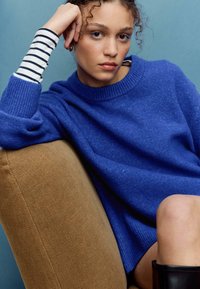 Kraljevsko modri ohlapni pulover z rebrastimi manšetami, kombiniran s črno-belimi črtastimi dolgimi rokavi spodaj. Ozadje rjave sedežne garniture.