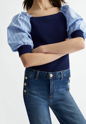 Femme bras croisés portant un haut en maille bleu marine avec des manches bouffantes rayées bleues et un jean bleu taille haute avec des boutons dorés sur les poches.