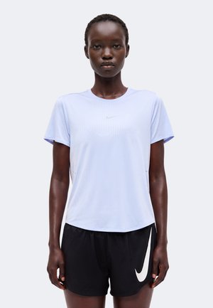 Kvinde iført en lyseblå sportst-shirt med korte ærmer og sorte atletikshorts med et hvidt Nike-logo, stående mod en ensfarvet baggrund.