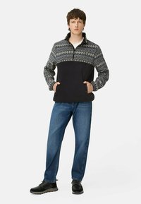 Sort og mønstret pullover med lynlås krave, kombineret med blå jeans. Pulloveren har et struktureret strikdesign på toppen. Sorte sko.