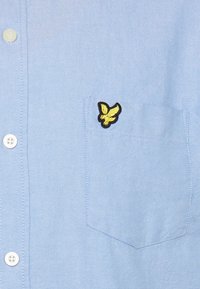 Camisa azul-claro de botões feita de tecido com uma textura subtil, apresentando um logótipo amarelo bordado de um pássaro no bolso do peito.