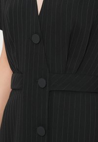 Μαύρο φόρεμα μπλέιζερ χωρίς μανίκια με ρίγες, τρία μεγάλα ματ μαύρα κουμπιά και ζωνάκι από το ίδιο ύφασμα στη μέση.