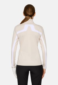 Maglietta a maniche lunghe, aderente, in tessuto beige con accenti riflettenti bianchi lungo la schiena e le maniche. Texture liscia, design con colletto alto.