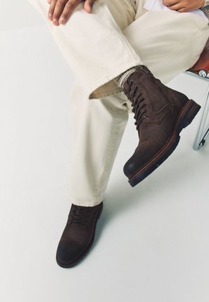 Osoba sediaca na stoličke so skríženými nohami, oblečená v svetlo béžových nohaviciach a tmavohnedých kožených šnurovacích topánkach s viditeľným šitím.