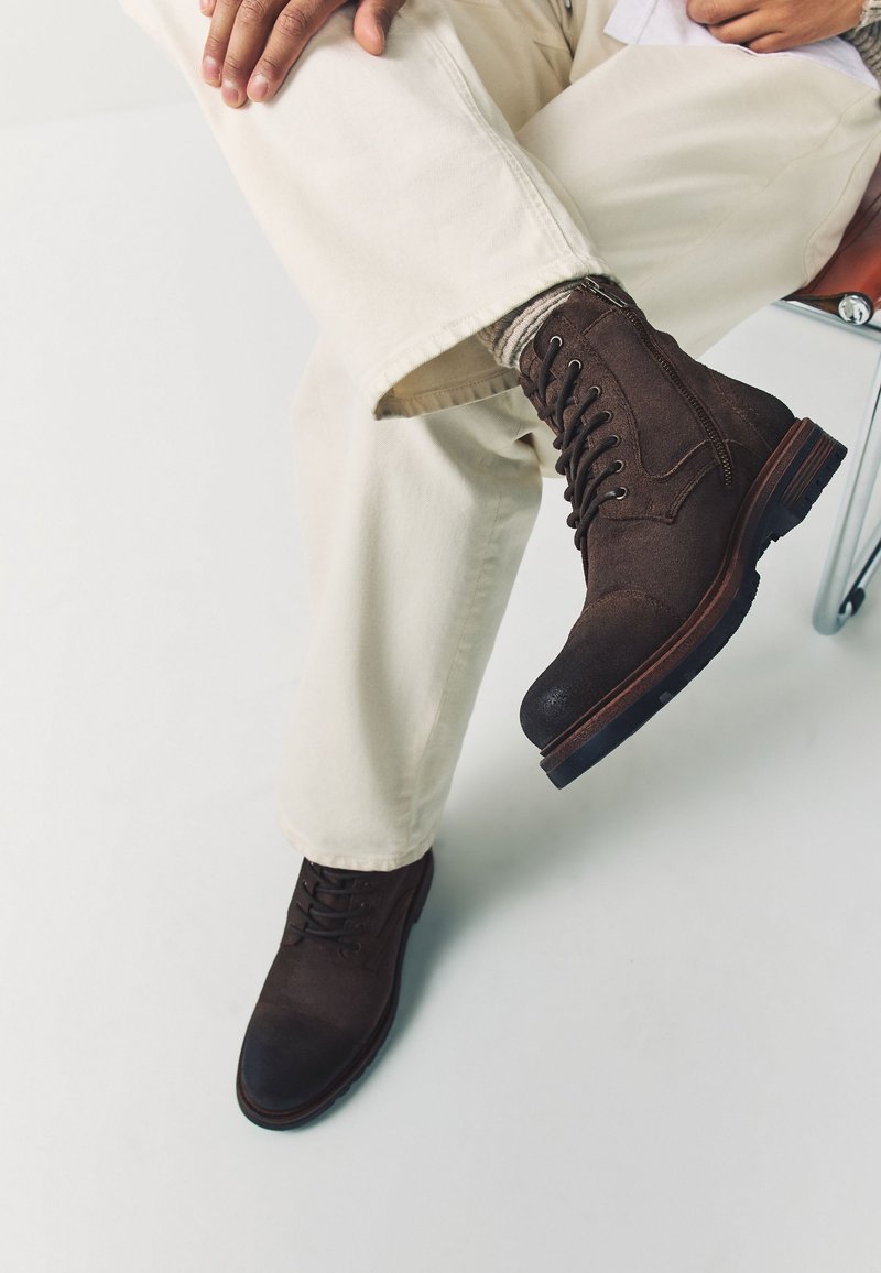 Persona sentada en silla cruzando las piernas, vistiendo pantalones beige claro y botas de cuero marrón oscuro con cordones y costuras visibles.