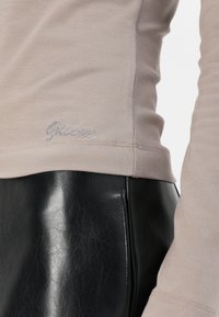 Tricou bej cu mâneci lungi, cu un design ajustat, având o broderie argintie "Guess"; asortat cu pantaloni din piele negri.