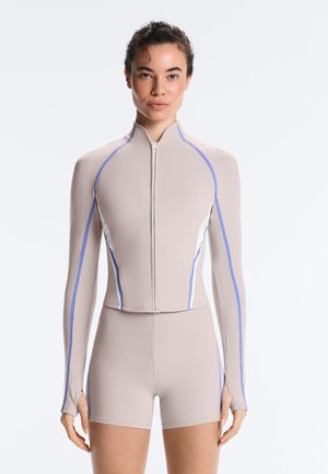 Donna che indossa una giacca beige aderente con zip, con strisce laterali blu e bianche e pantaloncini a vita alta abbinati su uno sfondo semplice.