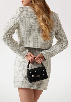 Borsa nera decorata con fiori e dettagli in perle, forma rettangolare e manico corto, tenuta dietro una persona che indossa un abito bianco texture.