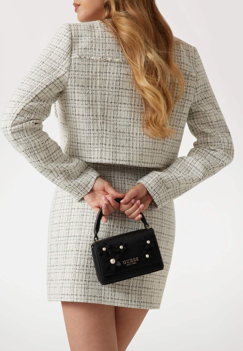 Black floral-embellished handbag with pearl accents, rectangular shape, and short handle, held behind a person wearing a textured white outfit.