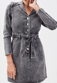 Robe en denim gris avec col, boutons sur le devant, manches longues, et ceinture nouée à la taille. Le tissu présente une finition texturée avec des coutures contrastantes.