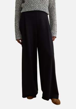 Une personne porte un pantalon large noir, un pull en maille gris et des chaussures en daim marron, debout devant un fond blanc.