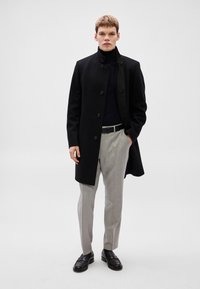 Casaco de lã preto com gola alta e fecho de botões, usado por cima de uma camisola de gola alta azul-marinho. Calças cinza claro e mocassins pretos completam o conjunto.