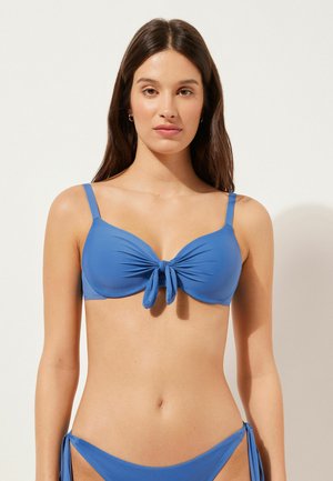Blå bikinitopp med knutdetalj i mitten, justerbara axelremmar och slät yta. Utformad för en åtsittande look.