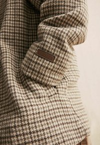 Brun og creme houndstooth mønsteret beklædningsgenstand lavet af et tekstureret uldlignende materiale. Har et læderlabel syet på ærmet.
