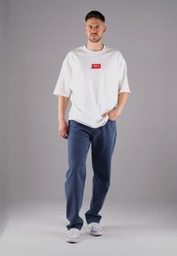 Weißes, übergroßes T-Shirt mit rotem Logo, kombiniert mit blauen Jeans und weißen Sneakers. Weicher Stoff, lockere Passform und einfaches Design.