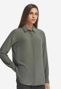 Camicia a maniche lunghe in verde oliva spento. Materiale liscio e setoso con colletto classico. Presenta una chiusura a bottoni sul davanti e una tasca laterale.