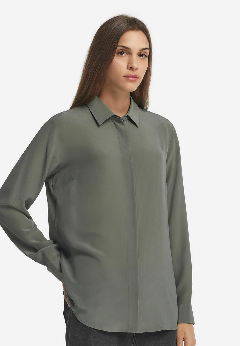 Langarmshirt in gedecktem Olivgrün. Glattes, seidiges Material mit einem klassischen Kragen. Verfügt über eine durchgehende Knopfleiste und eine seitliche Tasche.