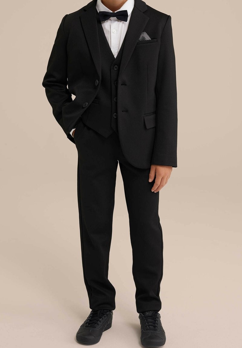 Svart tre-delad kostym med texturerat tyg, inkluderar kavaj, väst och byxor. Vit skjorta och svart fluga, bärs med svarta sneakers.