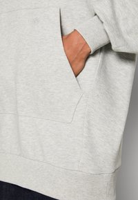 Sudadera gris claro en mezcla de algodón, con un bolsillo frontal, puños acanalados y un pequeño detalle de logotipo en el pecho. Textura suave.