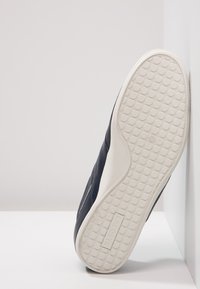 Suela de zapato mirando hacia adelante con patrón de agarre circular y logo de Lacoste embellecido en suela de goma blanca sobre fondo blanco.