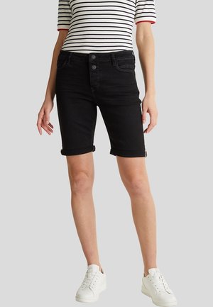 Kvinde iført sorte knælange shorts med opkrogede kanter, en hvid og sort stribet kortærmet skjorte og hvide sneakers, stående på en grå baggrund.