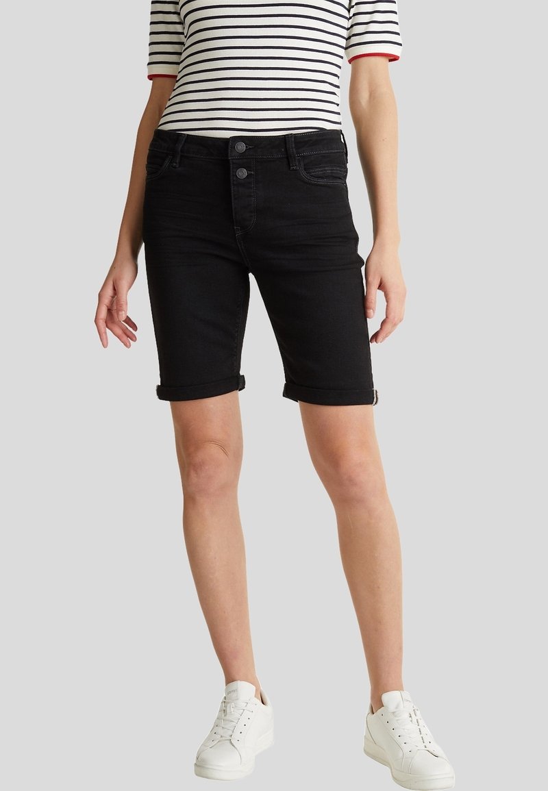Femme portant un short noir à ourlet roulé jusqu'aux genoux, un t-shirt à manches courtes rayé blanc et noir et des baskets blanches, debout sur un fond gris.
