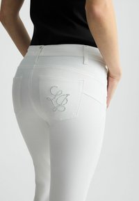 Valged denim teksased, millel on kitsas lõige, kaks tagumist taskut ja dekoratiivne logo, millel on sädelevad kristallid.