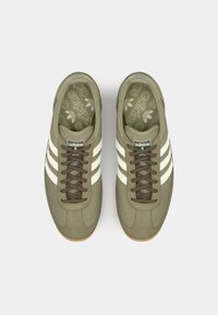Olive green sneakers featuring mesh and suede materials, three white stripes, brown laces, and a patterned insole with logo accents.