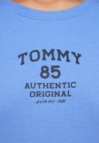 Zils ribots auduma tops bez piedurknēm ar melnu drukātu dizainu, kurā rakstīts "TOMMY 85 AUTHENTIC ORIGINAL since 1985." Apaļa kakla līnija.