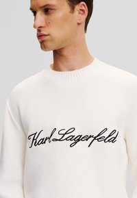 Baltas megztinis su raišteliais, turintis juodą siuvinėtą "Karl Lagerfeld" logotipą per krūtinę.