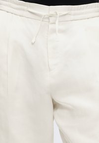 Κοντινό πλάνο λευκού παντελονιού με ορατό κορδόνι στη μέση και λεπτομέρεια μπροστινής ραφής σε άτομο που φοράει μαύρο τοπ.