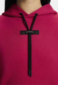 Fuchsiová mikina s kapucňou a šnúrkovým uzáverom, s čiernou etikettou s nápisom "ŠVAJČIARSKE INŽINIERSTVO." Hladká textúra, jednoduchý dizajn.