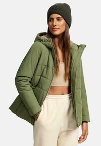 Olijfgroene pufferjack met capuchon, met een gewatteerde structuur en ritsluiting. Draagt over een beige cropped top en crèmekleurige sweatpants.