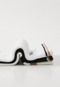 Des chaussettes rayées blanches, noires et marron pliées et empilées sur une surface blanche avec une forme ondulée.