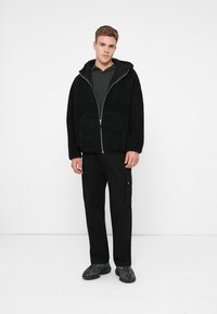 Casaco de fleece preto com fecho e capuz, duas bolsos frontais, combinado com uma camisa cinza escura e calças cargo pretas, usado com ténis pretos.