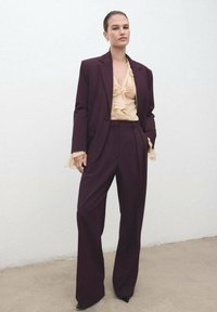 Traje burdeos oscuro con blazer ajustado y pantalones de pierna ancha. Blusa beige con volantes debajo. Tejido texturizado, silueta afilada, tacones de punta.