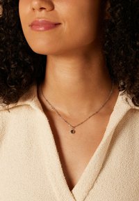 Zilveren ketting met een kleine groene hanger, gedragen op een beige textuur kledingstuk, rustend op het sleutelbeen.